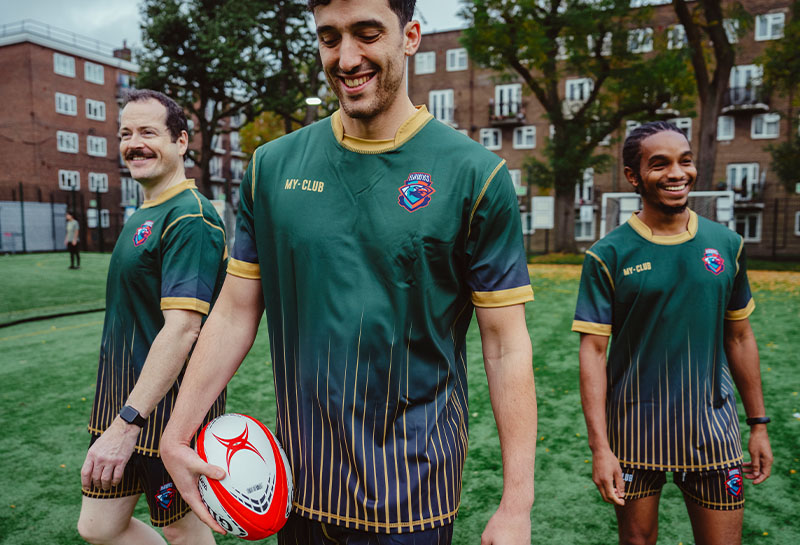 Three rugby players in green and gold uniforms stand on a grass field. One holds a red and white rugby ball.