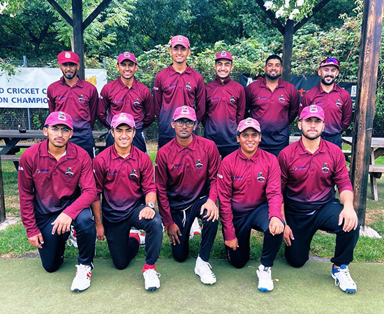 A cricket team poses for a group photo in matching maroon uniforms and caps outdoors.